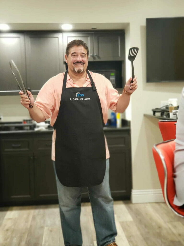 Dr. Alva wearing apron and holding kitchen utensils